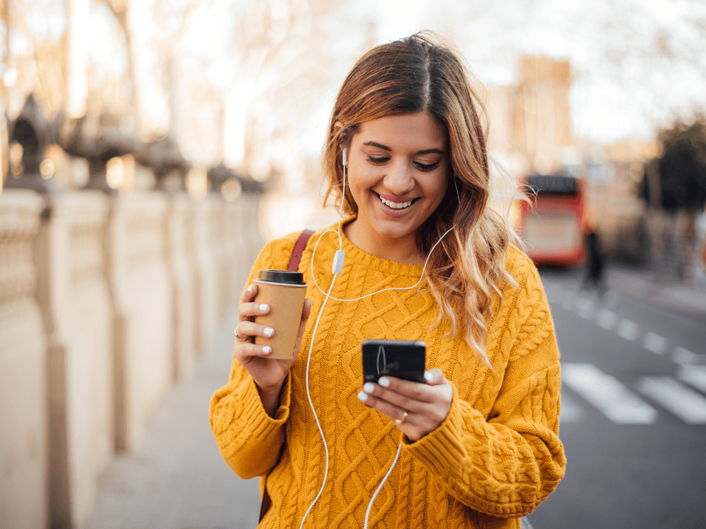 Happy woman walking through london with refurbished phone, coffee and earphones